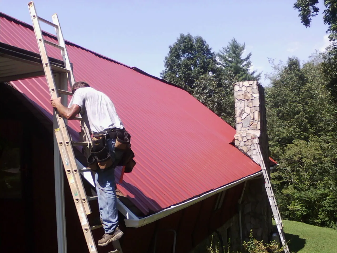 Roofer on a ladder beside a red metal roof and stone chimney — Soffit & Fascia Repair by Roofing Brothers in Sheakleyville