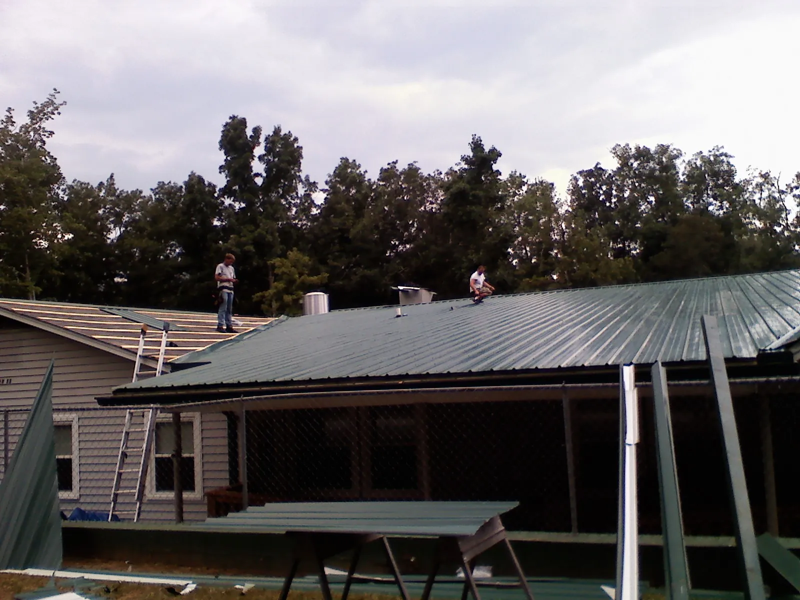 Two roofers installing green metal panels above a covered porch in Sheakleyville